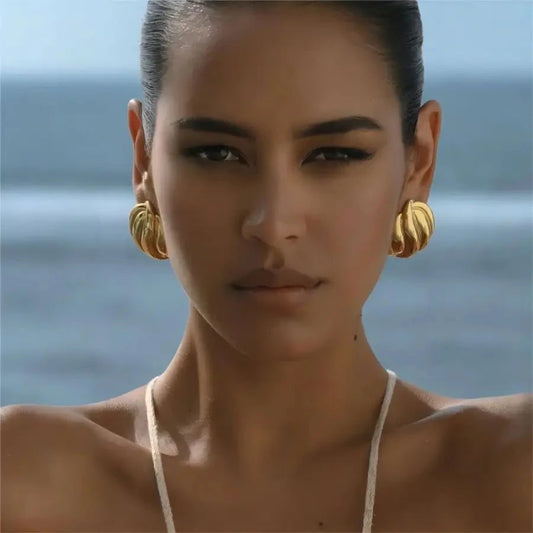 Woman with a bun wearing the Blair Gold Earrings against a blurred ocean backdrop, showcasing vacation-ready style and waterproof quality.