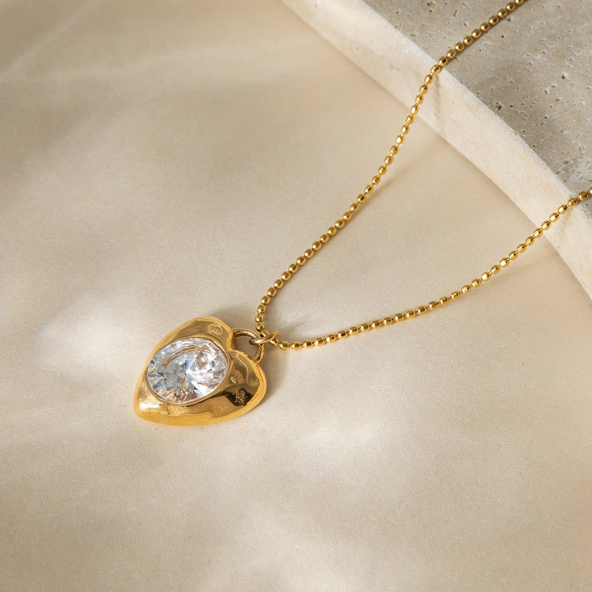 Flat lay of Bianca Crystal Heart Pendant Necklace—radiant crystal heart on gold-plated stainless steel chain.” Gold necklace with a heart-shaped pendant featuring a clear gemstone on a beige surface.
