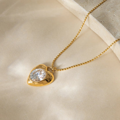 Flat lay of Bianca Crystal Heart Pendant Necklace—radiant crystal heart on gold-plated stainless steel chain.” Gold necklace with a heart-shaped pendant featuring a clear gemstone on a beige surface.