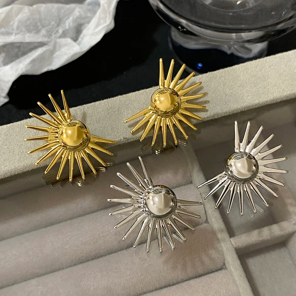A pair of bold sun-patterned stud earrings in gold and silver colors displayed on a grey surface.