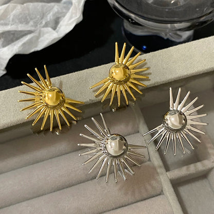A pair of bold sun-patterned stud earrings in gold and silver colors displayed on a grey surface.