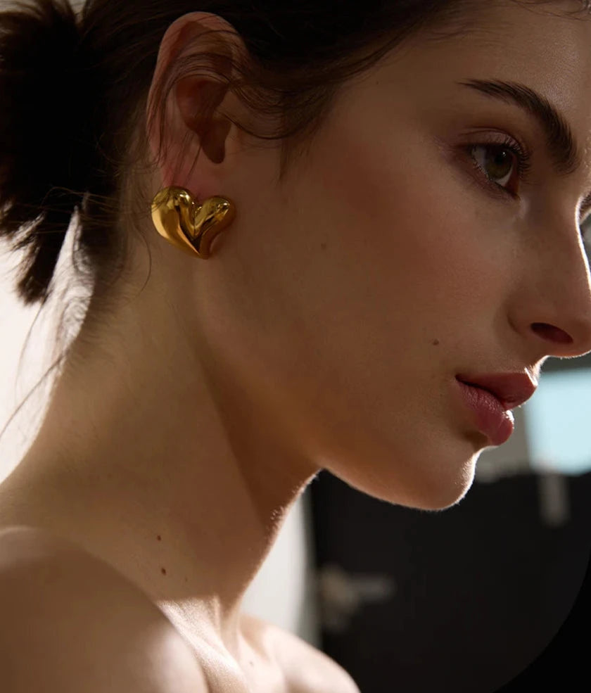 Model wearing chunky gold heart-shaped earrings.